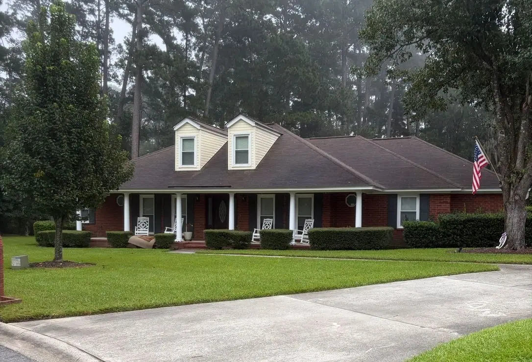 Before roof soft washing: red brick ranch house with dormers, a front porch and a flag, roof dark with algae streaks