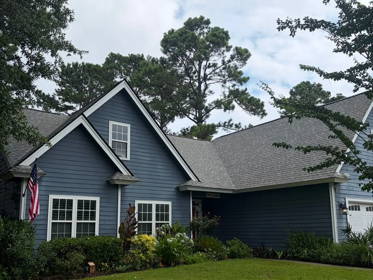 After roof soft washing: the same blue lap-siding house with a gray shingle roof under pine trees, roof clean