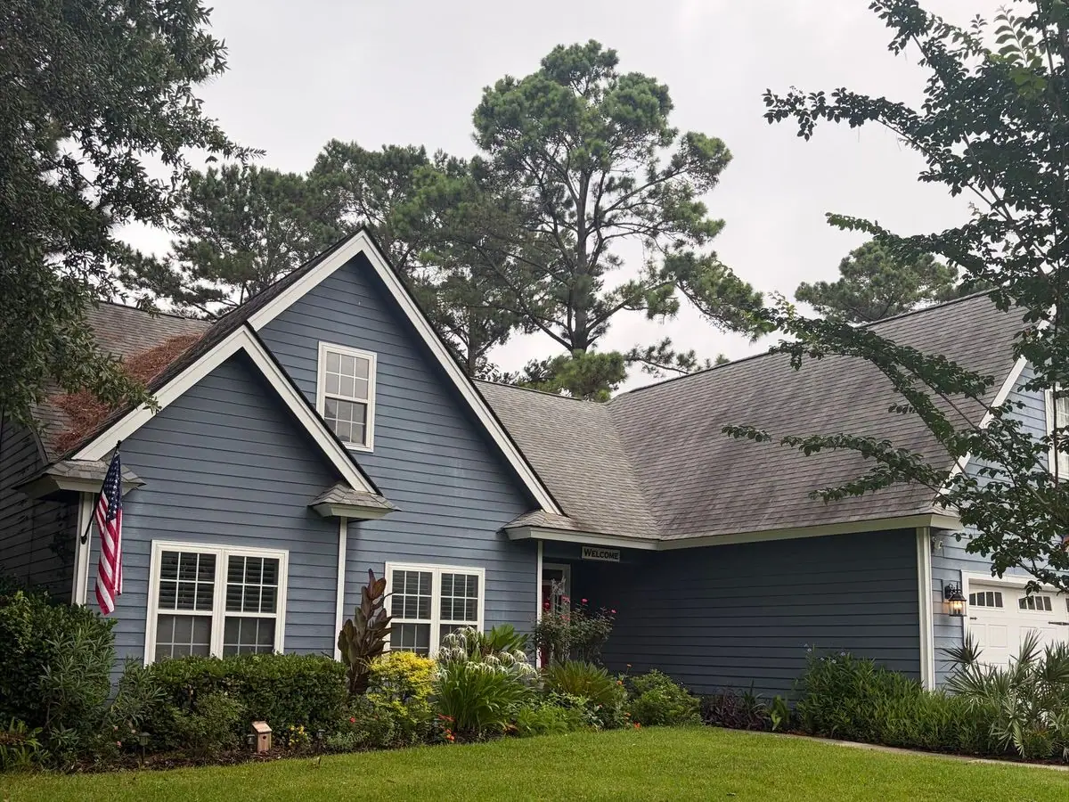 Before roof soft washing: blue lap-siding house with a gray shingle roof under pine trees, roof dark with algae streaks